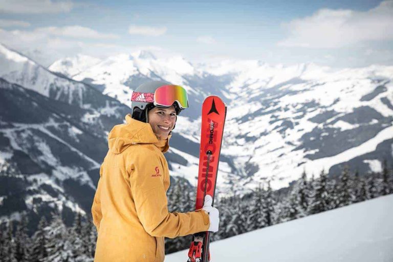 Skifahrerin mit Sonnenbrille und Ski vor schneebedeckten Alpen im Hintergrund.